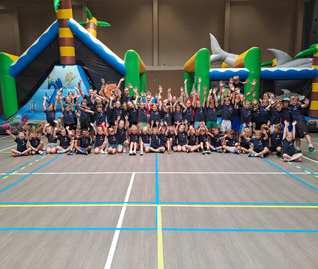 Grote groep kinderen in Sportiek-t-shirts poseert al juichend tijdens een Sportiek sportkamp, met springkastelen op de achtergrond.